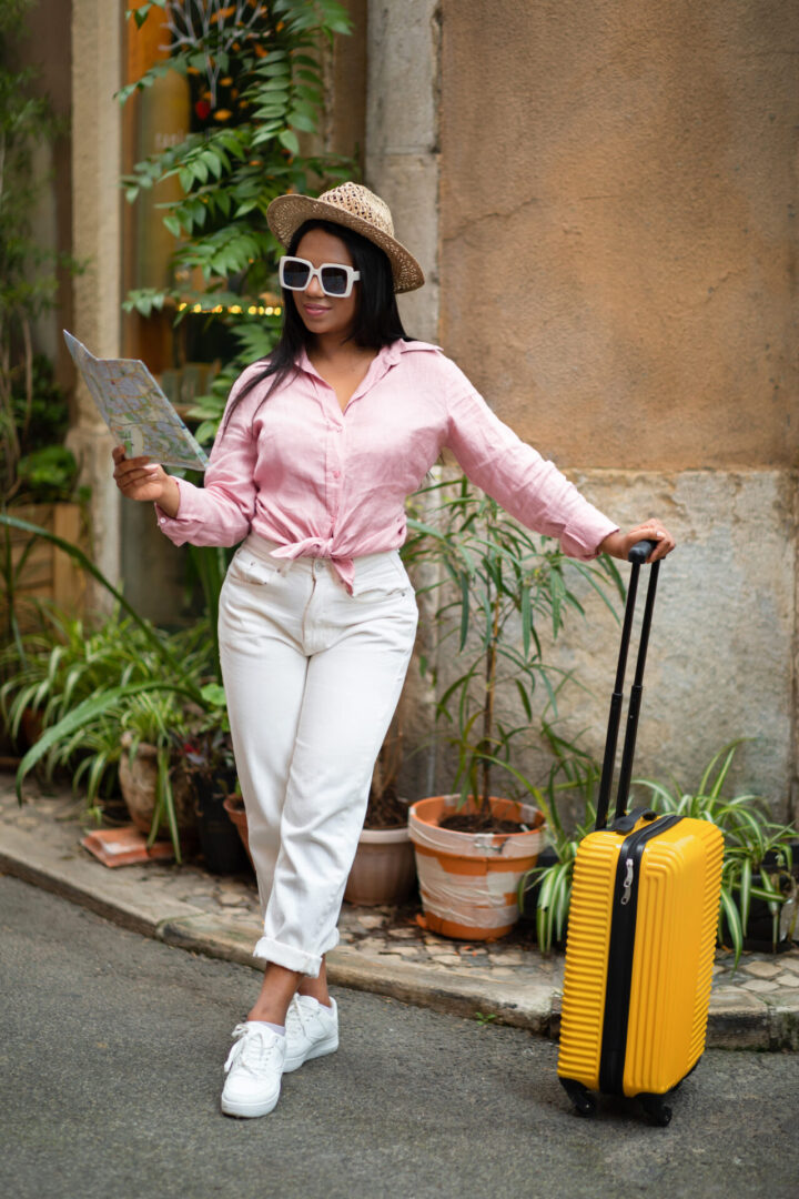 Glad young black lady in casual sunglasses with suitcase looking at map, enjoy trip in new city, outside, vertical. Rest, vacation and travel summer, road to hotel, adventure